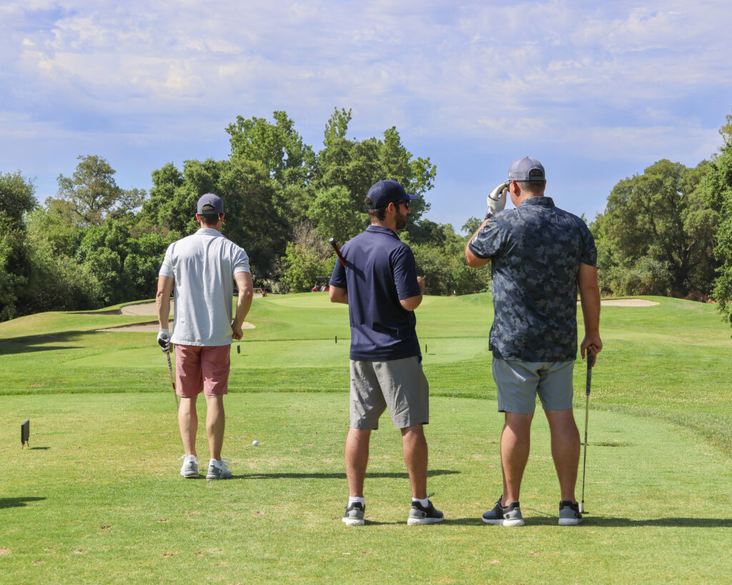 Three golfers stand on the tee box on the 16th hole on the MacKenzie course. One of them is preparing to tee off while the other two watch.