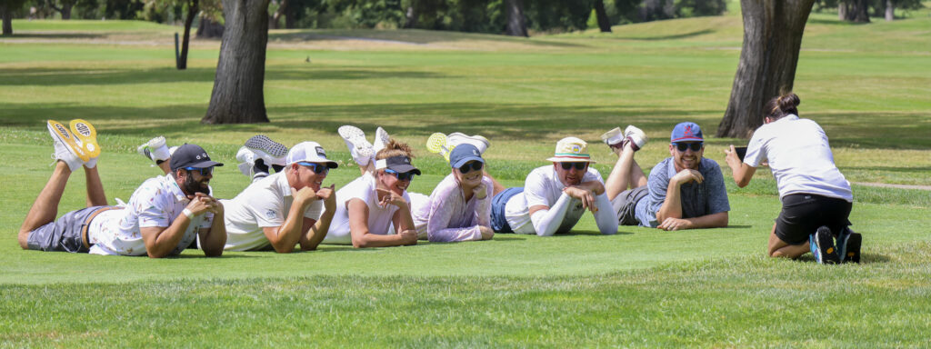 A group of 6 golfers posing for a photo. They are all laying down on their stomachs with their legs crossed and raised behind them. They are resting their head on the hands while another person takes their photo.