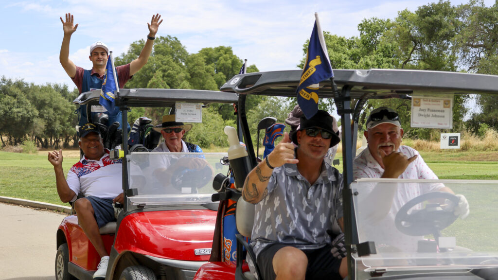 A group of golfers posing for a photo in their golf carts. The two in the front are making a 'shaka' hand sign while the other 3 people behind them have their hands raised.