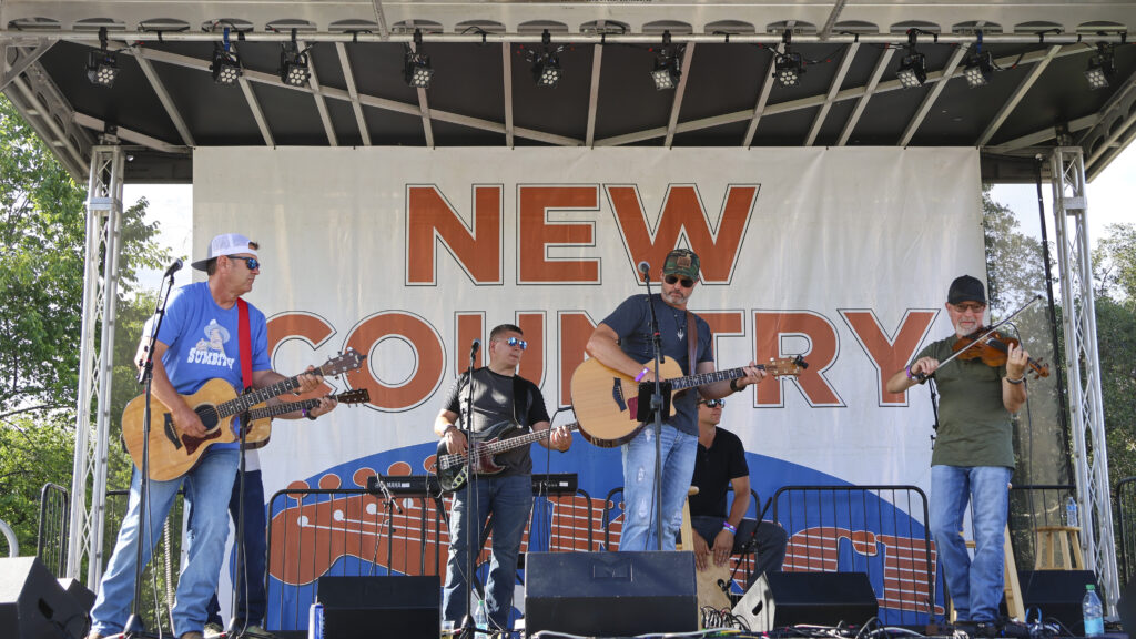 Cripple Creek Band on the main Golf & Guitars concert stage. Behind them is a large sign that reads 'NEW COUNTRY'.