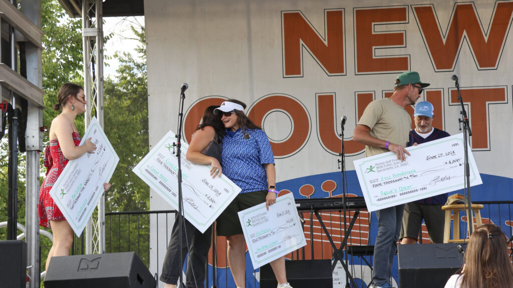 Mae Estes, Kennedy Scott, Erin Enderlin, and Eric Gunderson on stage with their giant checks for their charities. Erin and Kennedy are hugging while Mae and Eric are looking to the left side of the stage.