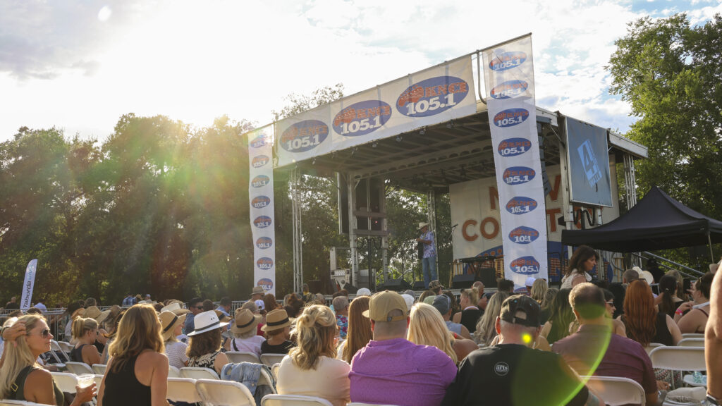 A wide-shot from the audience's perspective of the main show stage. The crowd is packed and the sun is peeking over the trees, causing a lens flare.