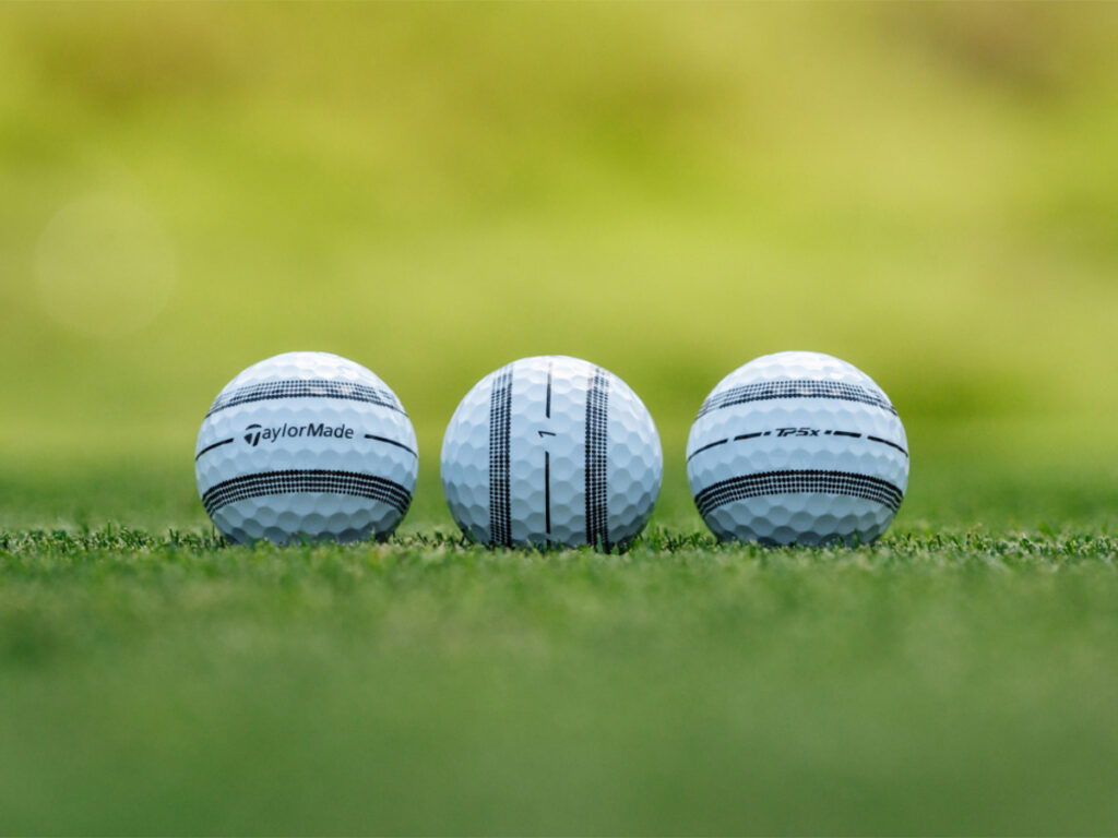 Three TaylorMade TP5x Stripe balls sit on grass, lined up so you can see different parts of the ball. You can see the stripe, the logo, and the ball number.