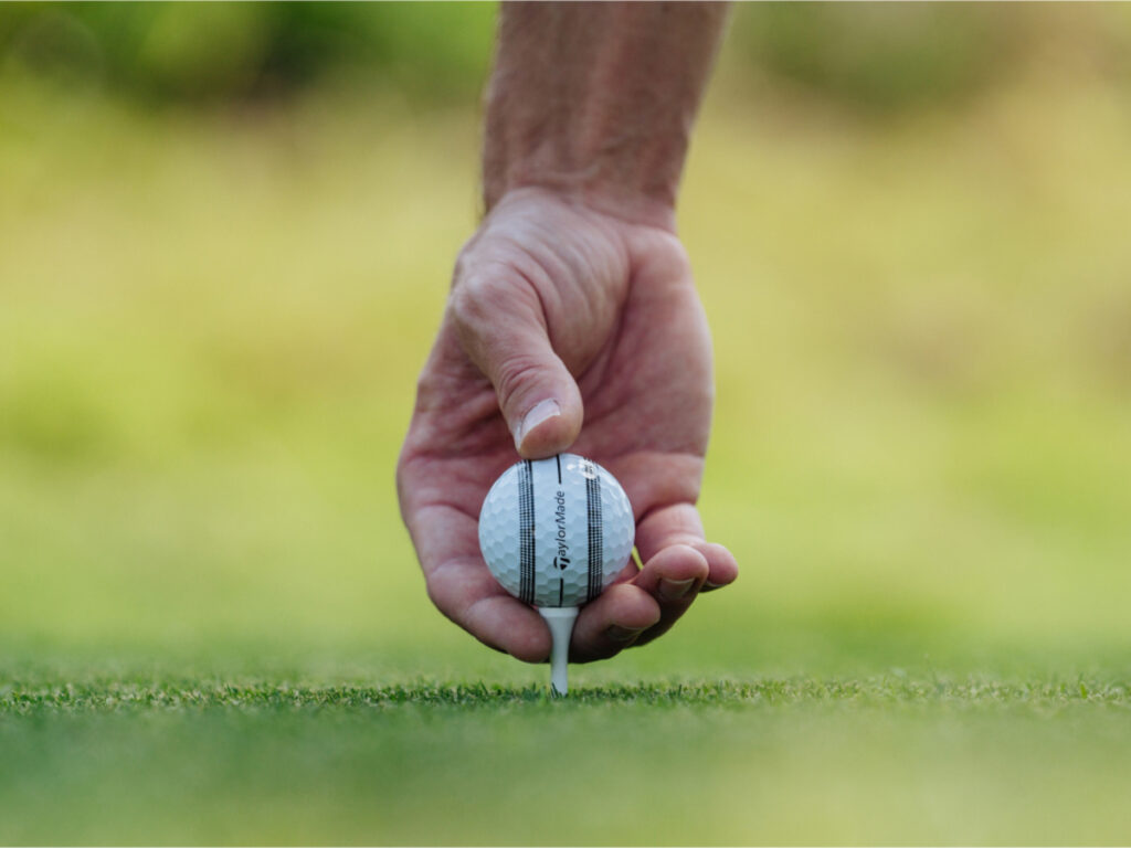 Close-up of a hand reaching down to place a TaylorMade TP5x Stripe ball on a golf tee.