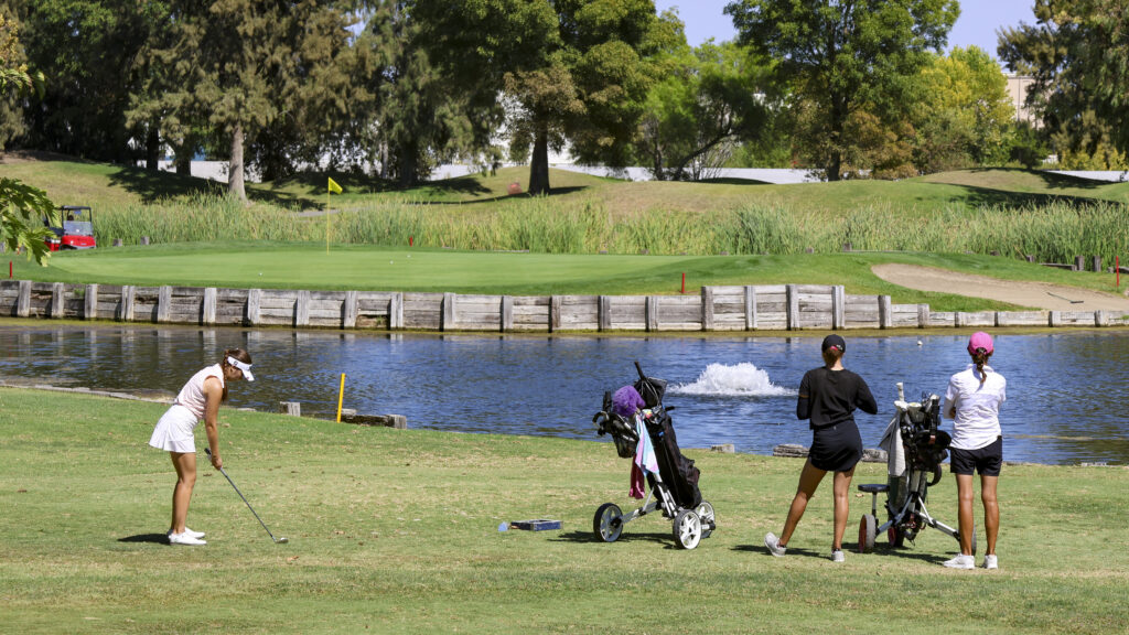 A wide-shot of the 17th tee at Bartley Cavanaugh. Sienna Lyford is preparing to tee off with the 17th green in the distance. Tori James is standing off to the side, watching Sienna.