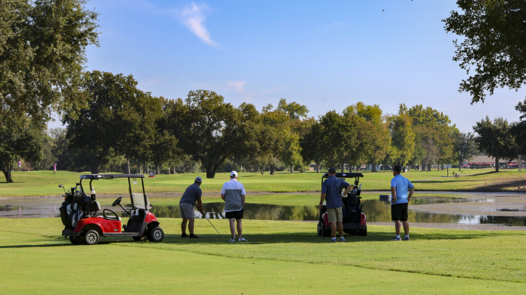 A group of 4 golfers stand around their 2 golf carts by the lake on the 4th hole. One of them is preparing to hit their ball.