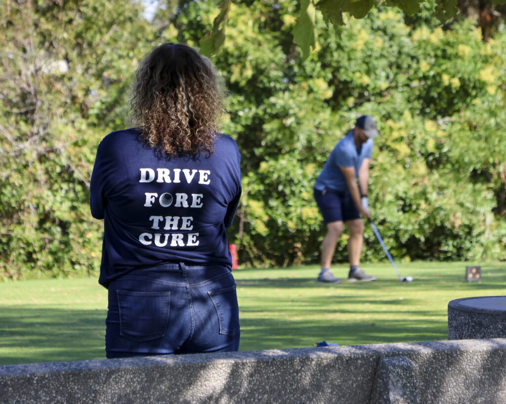 A woman watches as a man is preparing to hit his ball on the 14th hole. The woman is wearing a Jones'n for a Cure shirt that reads "Drive Fore the Cure".