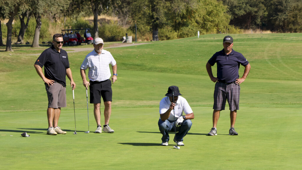 A group of 4 golfers standing on the #16 green. One of them is squatting on the ground, trying to read the green before making his putt.