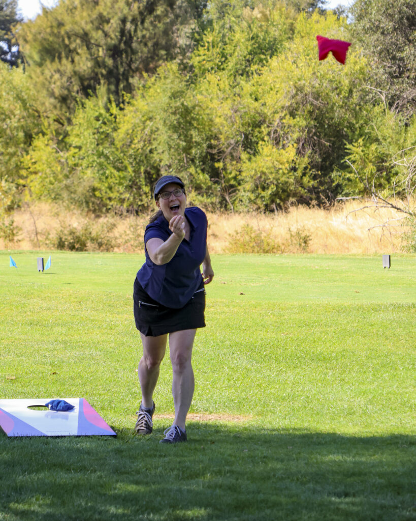 A female golfer is playing cornhole. She has just thrown her bag, hanging high in the air. Her mouth is open in excitement.