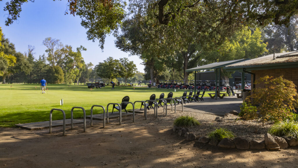 A wide shot of the William Land Golf Courses's practice putting greens and clubhouse. One person is putting on the green while a maintenance staff member is tending to the green. In the distance, golf carts are lined up for customers to take out.