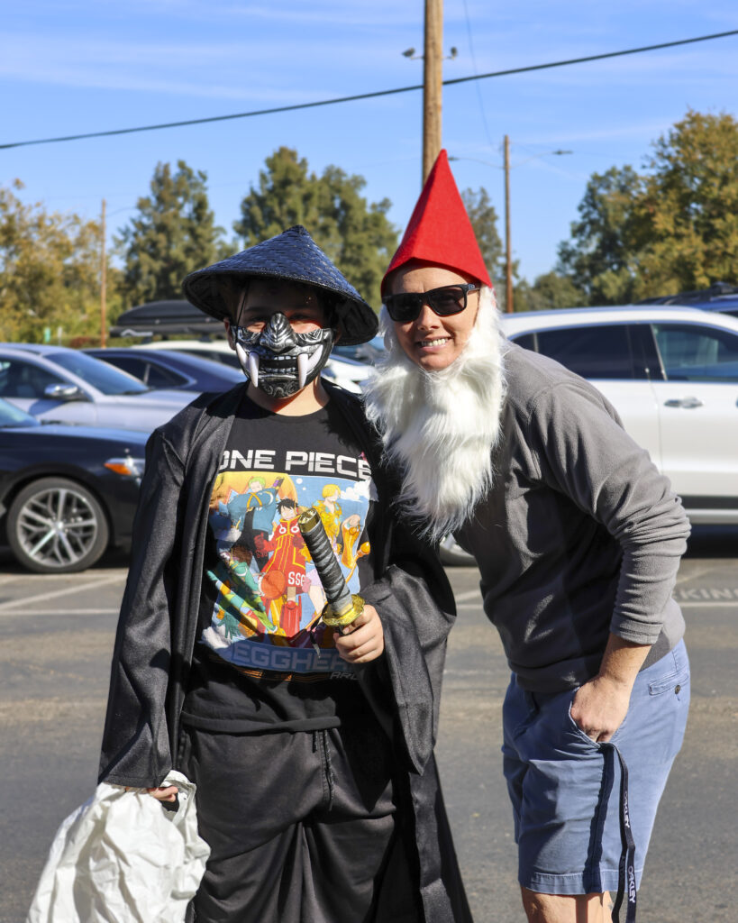 A teenager and their parent pose for a photo in the parking lot. The teenager is in a black outfit wearing an oni mask and a katana. The parent is wearing a red gnome hat and a large fake white beard.