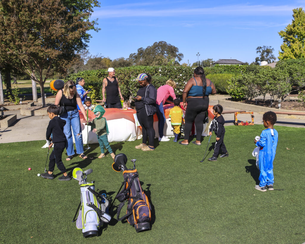 A large group of children and parents are gathered around a long table, coloring pictures and playing with golf clubs.