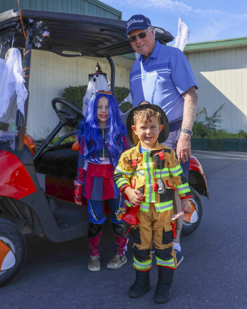 A grandfather poses with his two grandkids. The grandson is dressed like a fire fighter and the granddaughter is dressed like a superhero.