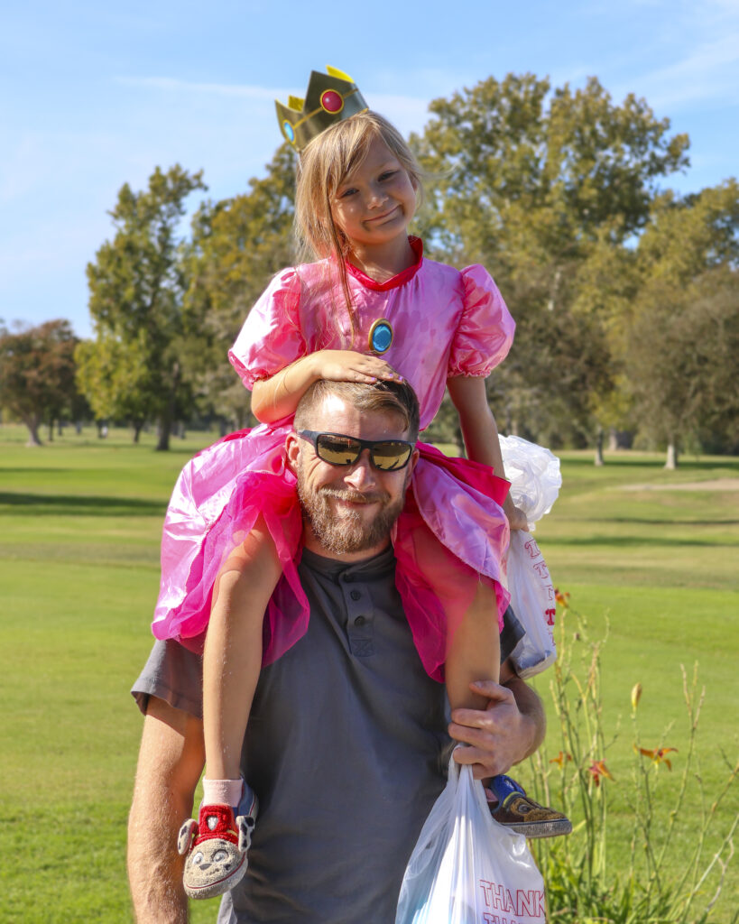 A father and daughter pose for a photo. The daughter is dressed like Princess Peach while sitting on her father's shoulders, who is dressed in normal clothing.