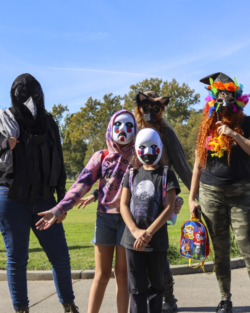 A large group of women pose for a photo, all wearing masks. Two are wearing white masks with black eyes and red cheeks while the others wear masquerade masks.