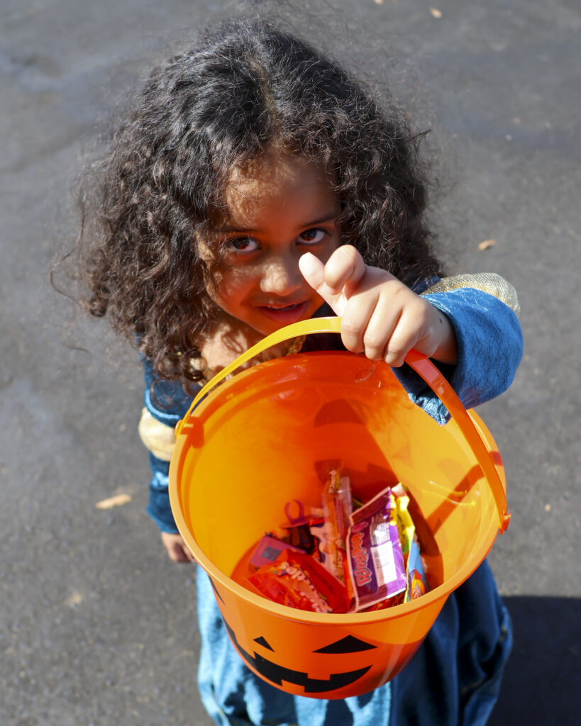 A child dressed like Meridia from Brave is holding up her trick-or-treat bucket full of candy to the camera.