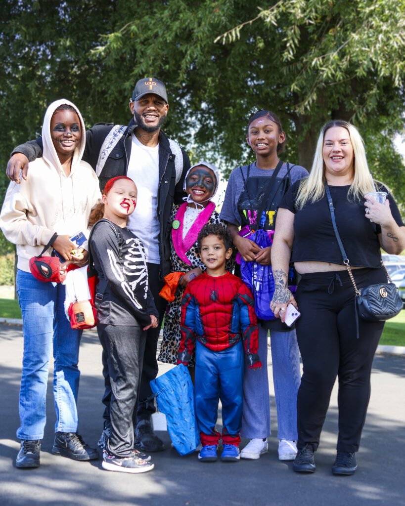 A large group of 7 is posing for a photo. One teenager is dressed as a zombie, another is a skeleton, and a young boy is dressed like Spiderman. 