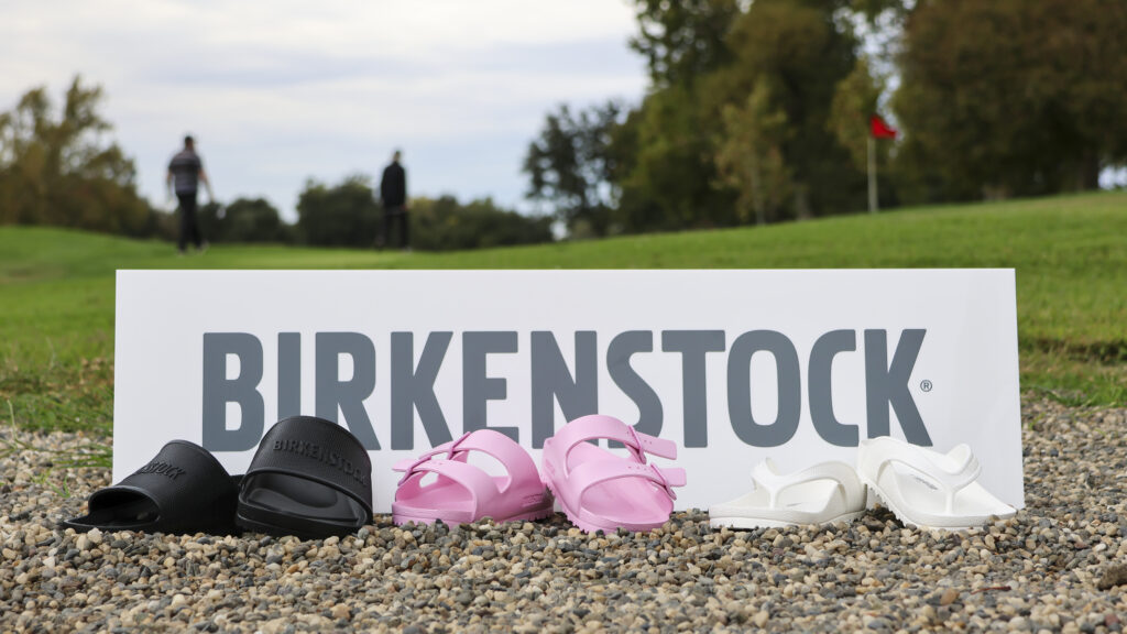 3 pairs of different style Birkenstock EVA sandals are lined up in front of a sign that says 'BIRKENSTOCK'. The leftmost pair is black, the middle pair is pink, and the rightmost pair is white. They are outside sitting on gravel, with a golf hole and two golfers behind them in the background.