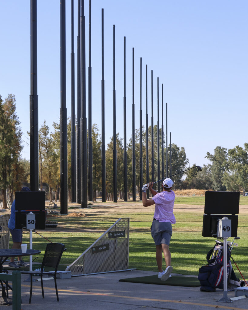 A man is hitting on the driving range. Behind him are the net pillars after being installed. Some upturned dirt can be see at the base of one pillar.