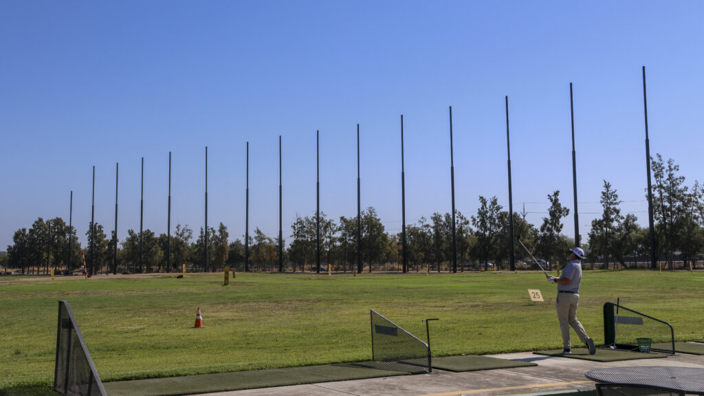 A wide shot of the range while a man hits onto the driving range. Beyond him in the distance the net pillars can be seen without the net installed.